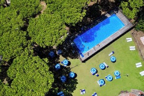Eine Vogelperspektive auf ein Schwimmbecken, umgeben von üppiger Vegetation, mit mehreren Liegestühlen und Sonnenschirmen im Poolbereich.