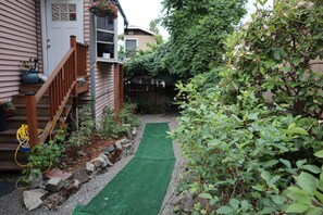 Un jardin à l’arrière d’une maison, avec un escalier en bois, un chemin en gazon synthétique vert et diverses plantes.