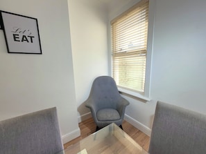 A dining area with a glass table, two grey chairs, a framed sign saying 'Let's EAT', and a window with blinds. - Family & Work Stay | Garden, Parking & Workspace (Southend-on-Sea)