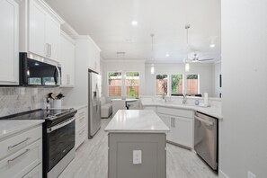 A modern kitchen with white cabinetry, a central island, stainless steel appliances, and a view of the backyard through large windows. - The Oak House - Love Street Collection 
Luxury New Build Near Love St. Park (Weatherford)