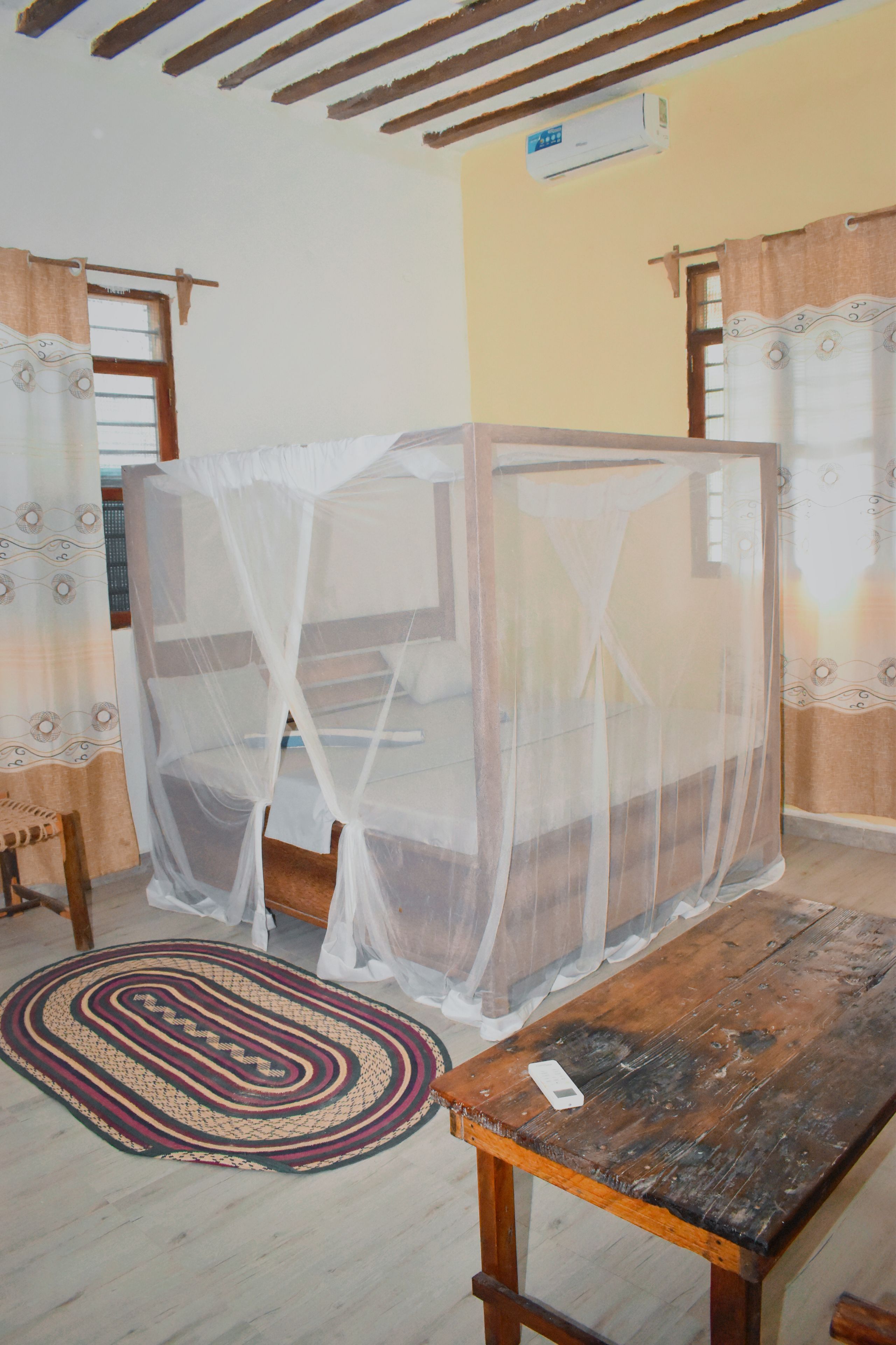 A room with a bed covered by a mosquito net, a wooden table, and a patterned rug.
