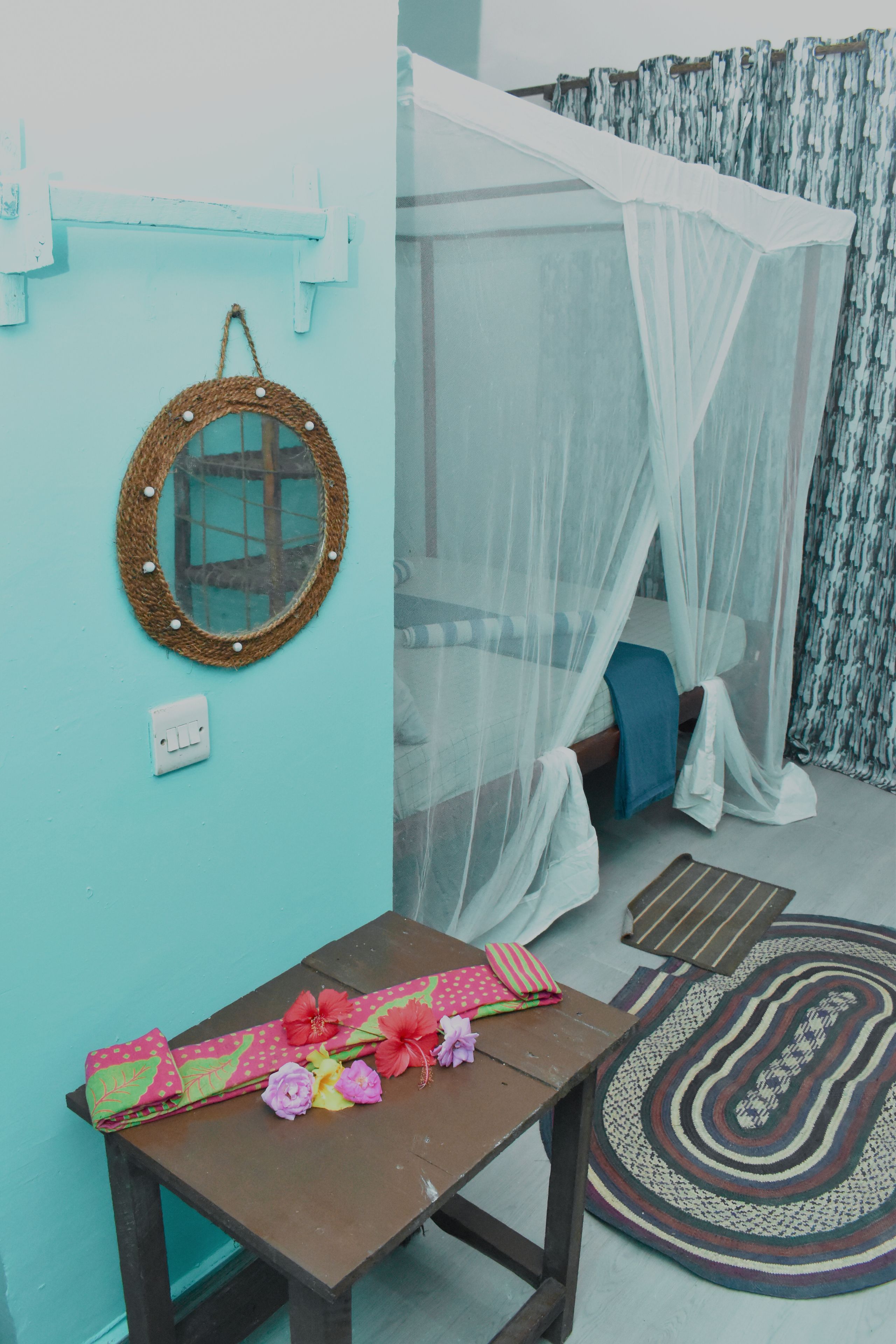 Una habitación con una cama bajo una mosquitera, un espejo redondo en la pared, una mesa con un mantel colorido y una alfombra estampada.