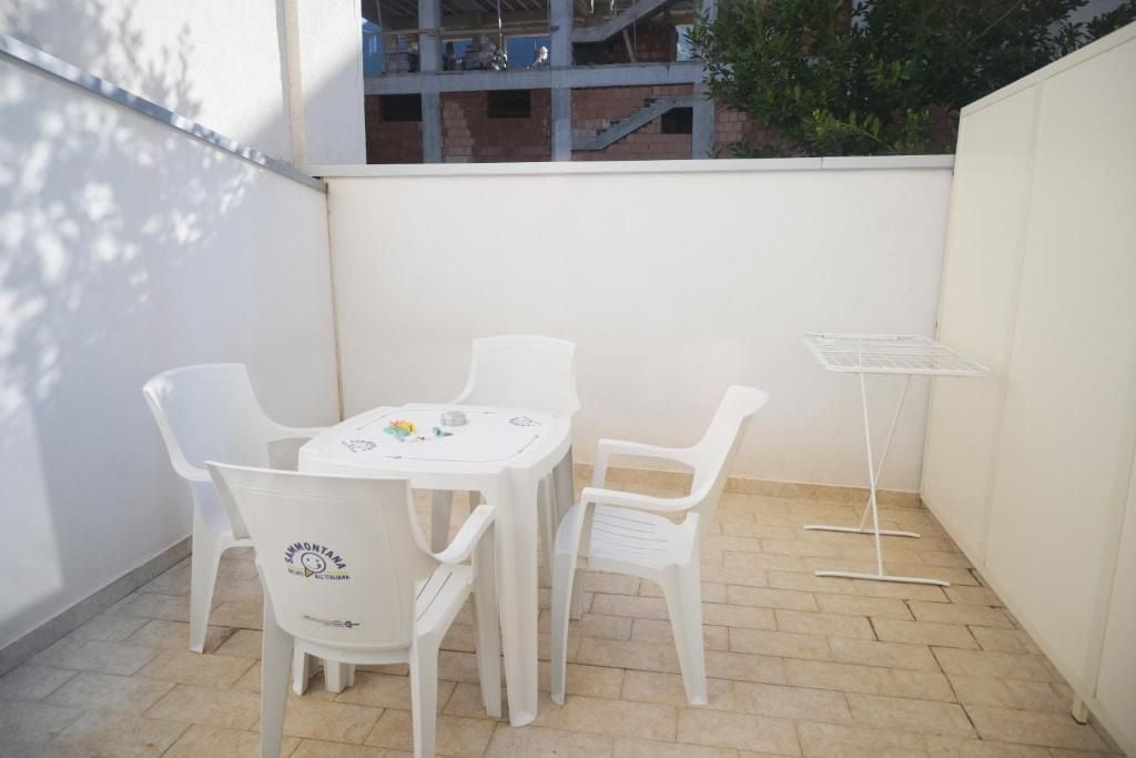 Une petite terrasse extérieure avec une table et des chaises blanches, un présentoir en métal blanc, et en arrière-plan, une vue sur un bâtiment en construction.