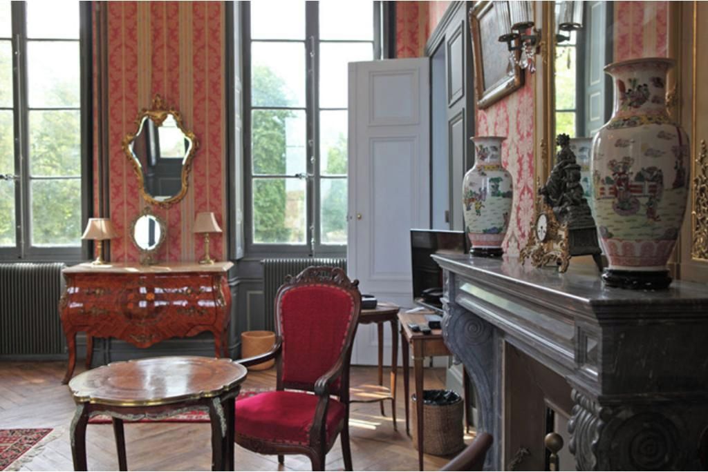 Une pièce avec une cheminée, un fauteuil rouge, une table en bois et un miroir.