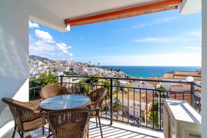 A balcony with wicker furniture and a glass table overlooking a coastal town. - Apartment in Nerja Near Burriana Beach (Nerja)
