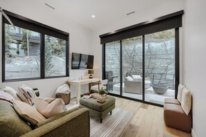 A modern living room with a large window, a flat-screen TV, a sofa, a coffee table, and a dining area with outdoor seating. - 1061 Lowell Avenue #4 (Park City)