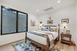 Una camera da letto con un'ampia finestra, un letto con lenzuola fantasia, una testiera in legno, un comodino con una lampada e opere d'arte appese al muro.