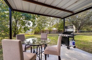 Un patio exterior con una mesa redonda de vidrio y cuatro sillas, una parrilla de barbacoa y un tanque de propano.