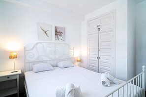 A white bedroom with a bed, two bedside lamps, a nightstand, a wardrobe, and two wall art pieces. - Capistrano Village Sundreams by Casasol (Nerja)