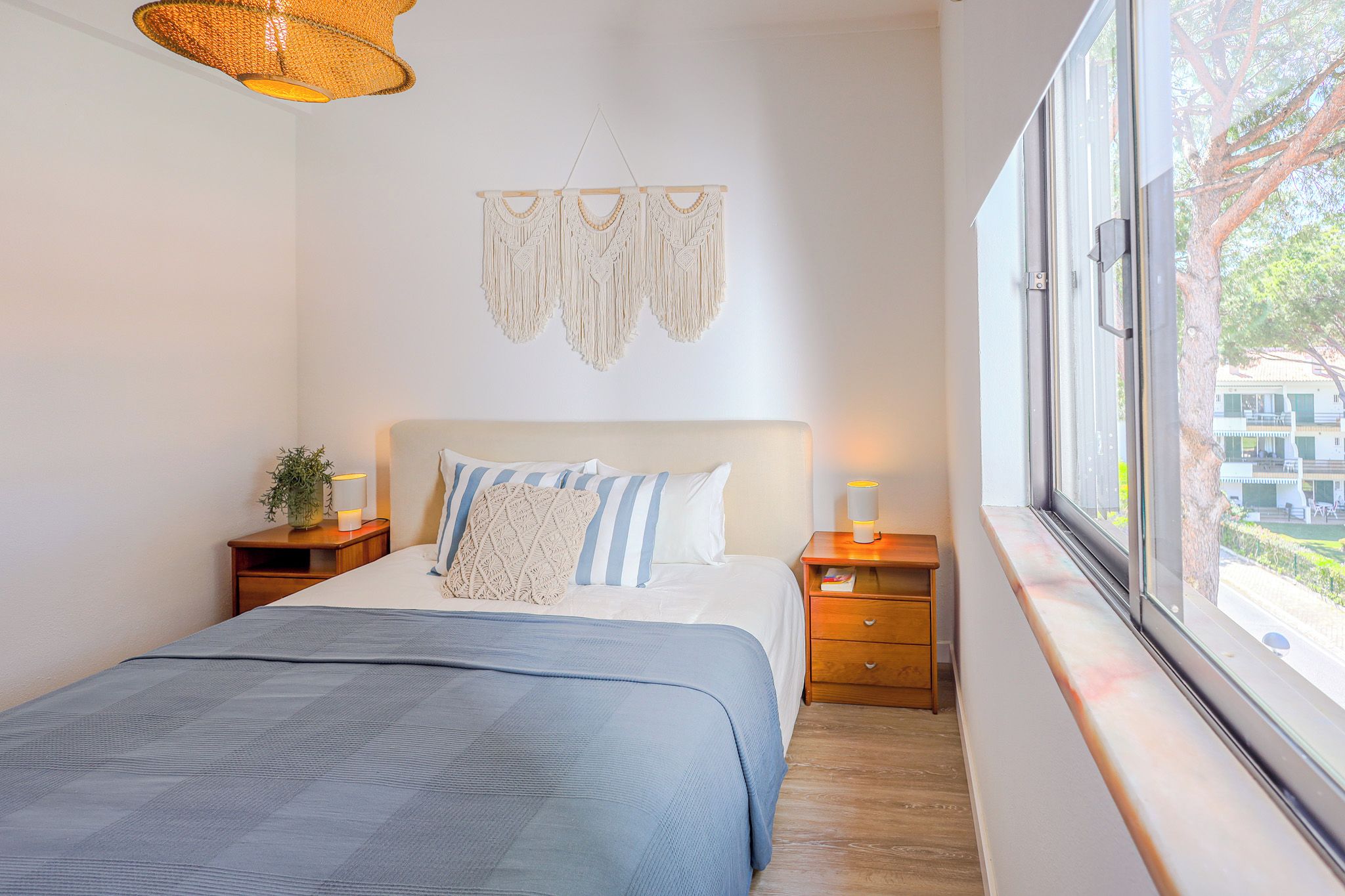 A bedroom with a bed, bedside tables, a macrame wall hanging, and a window with a view.