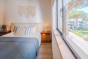 A bedroom with a bed, bedside tables, a macrame wall hanging, and a view of a building through a window. - Sun & Golf Vilamoura Boutique Apartment (Quarteira)