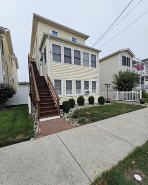 A two-story residential building with a wooden staircase leading to the entrance, a front yard with a white picket fence, and a clear sky. - 1 Bedroom/1 Bathroom Beach Getaway (Ocean City)