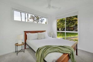 Ein Schlafzimmer mit einem großen Bett, einem Nachttisch, einer Lampe und Blick auf einen Garten durch ein großes Fenster.