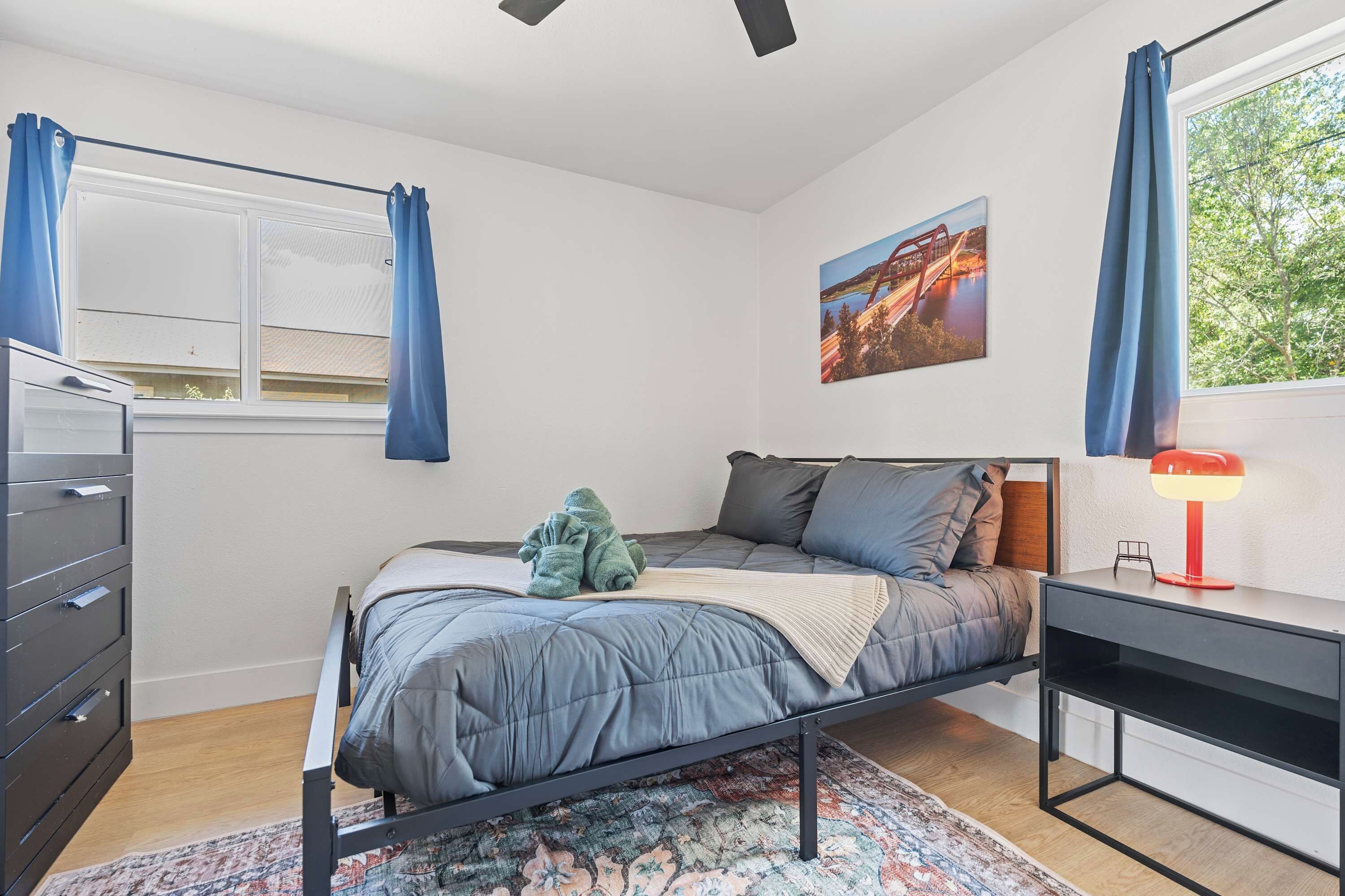 Un dormitorio con cama, escritorio, silla, cómoda, ventana con cortinas y un cuadro en la pared.