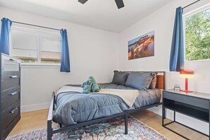 Un dormitorio con cama, escritorio, silla, cómoda, ventana con cortinas y un cuadro en la pared.