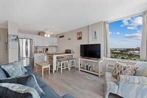 A modern living room with a large TV, a dining area, and a kitchen. - Estero Beach & Tennis 802c - Beach Sunrise (Fort Myers Beach)