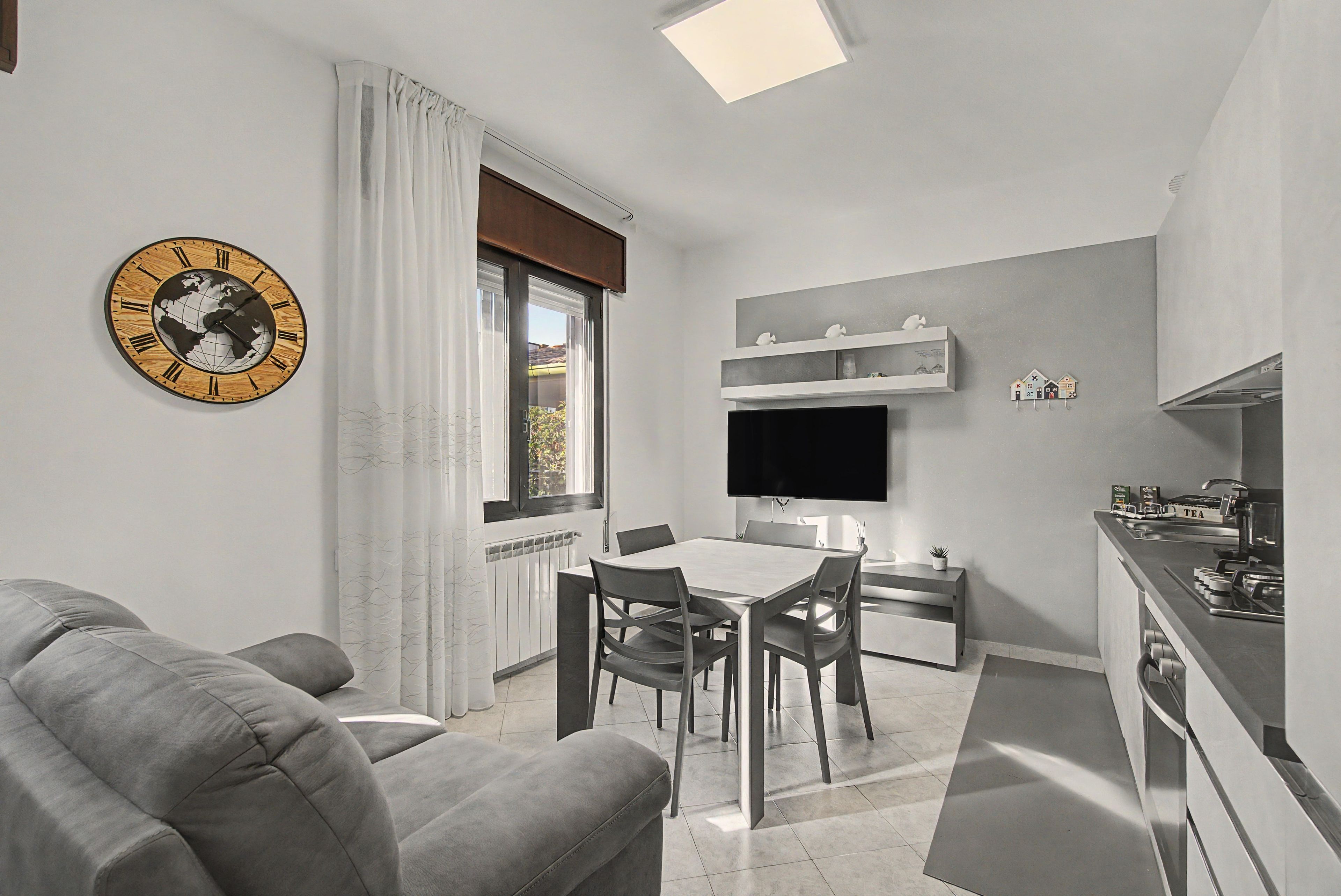 Een moderne woonkamer met een grijze bank, een eettafel met stoelen, een flatscreen televisie aan de muur en een keuken met een fornuis en spoelbak.