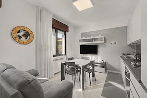 A modern living room with a grey sofa, a dining table with chairs, a flat-screen TV mounted on the wall, and a kitchen area with a stove and sink.
