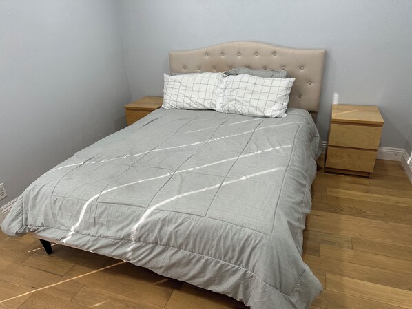 Uma cama com cabeceira estofada, um edredom cinza e duas mesas de cabeceira com tampos de madeira.