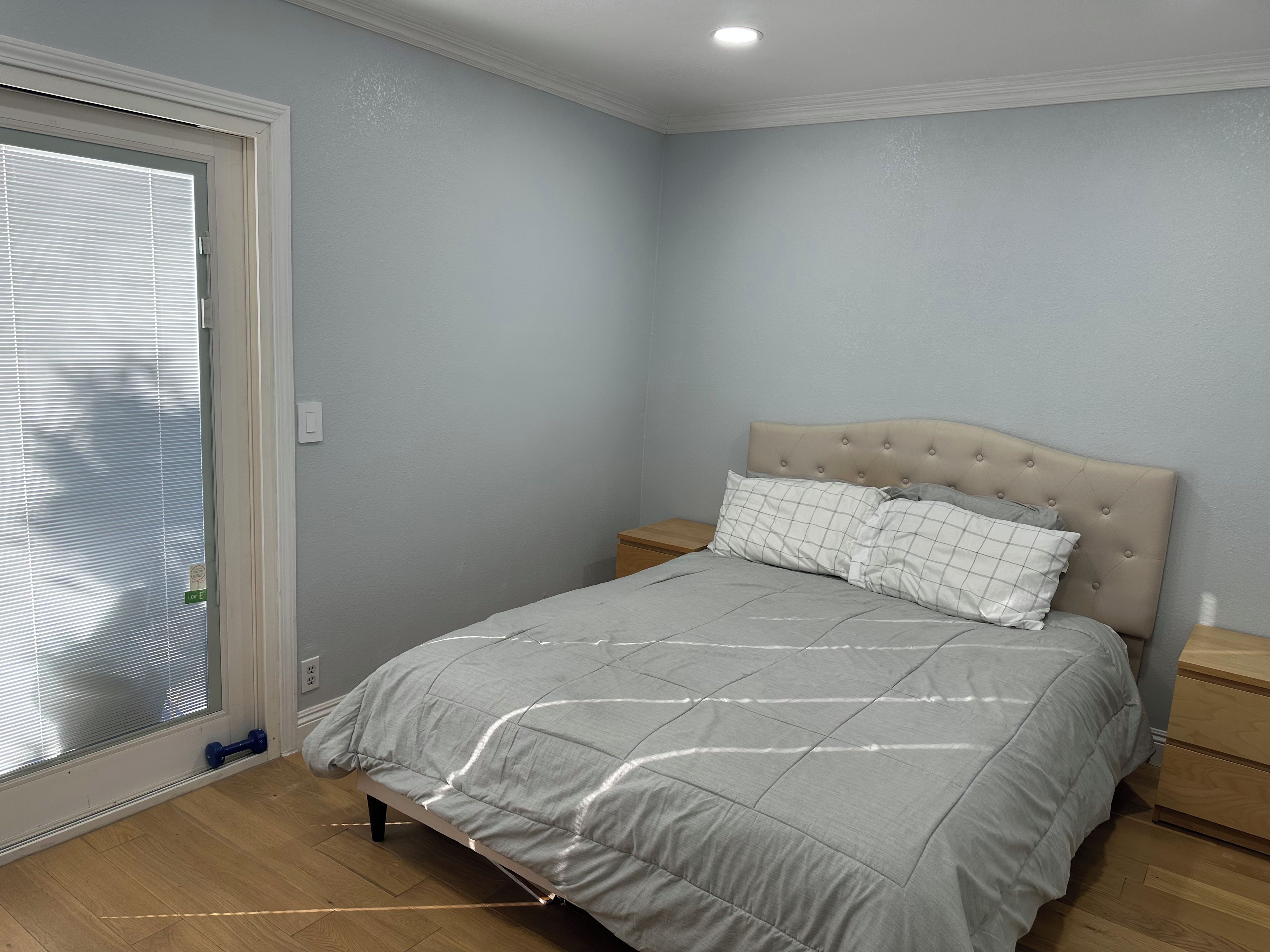 Een slaapkamer met een bed, nachtkastje, raam met zonwering en een deur.