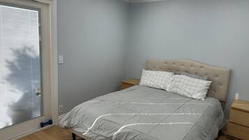 Een slaapkamer met een bed, nachtkastje, raam met zonwering en een deur.