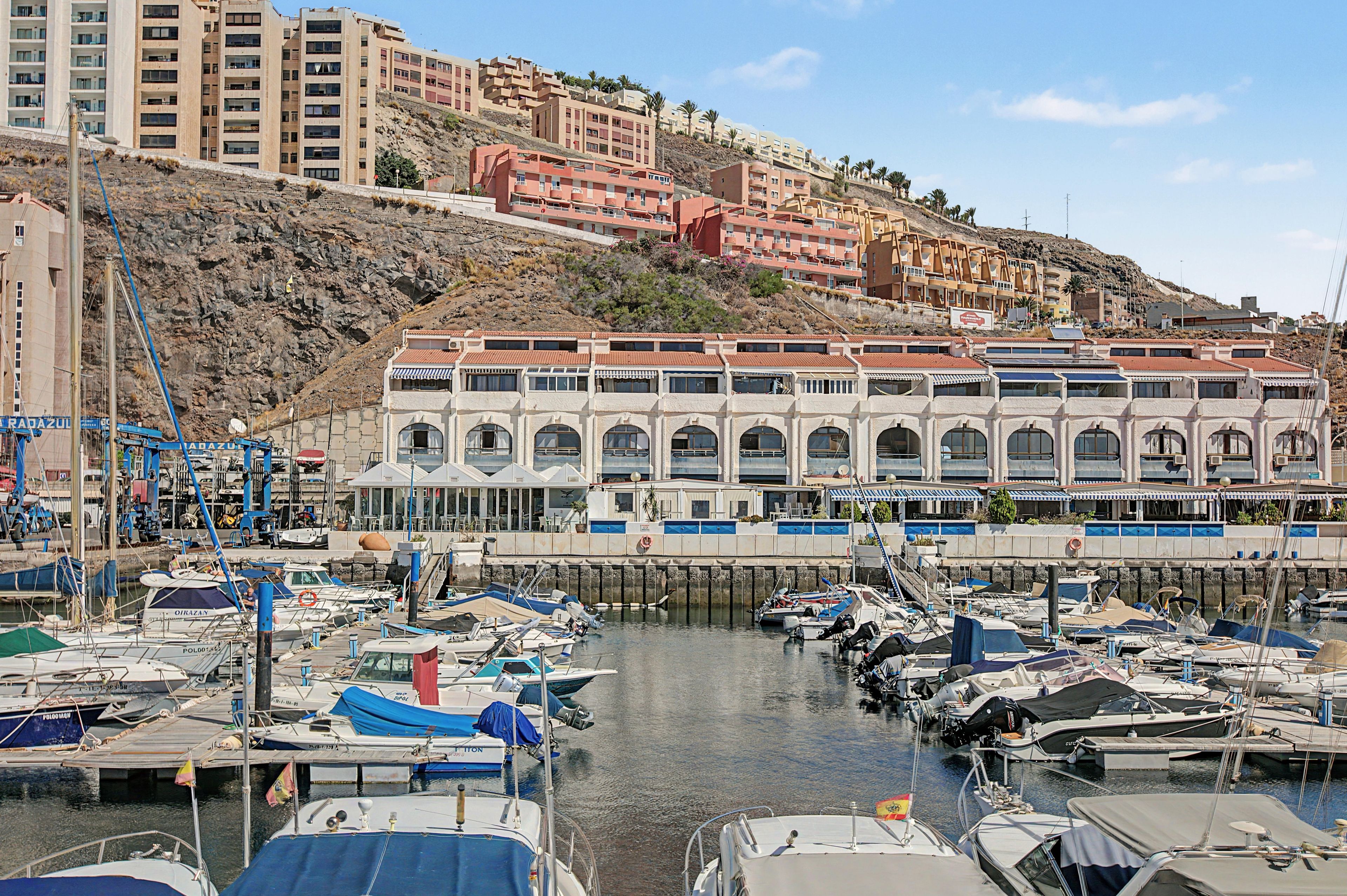 Un port de plaisance où sont amarrés de nombreux bateaux, un grand bâtiment à la façade blanche et aux fenêtres en arc de cercle, et un paysage vallonné en arrière-plan, parsemé de bâtiments résidentiels.
