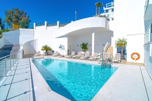 A swimming pool with clear blue water, surrounded by white lounge chairs and a white building with a curved roof. - Apartment in Nerja With Sea Views & Pool (Nerja)