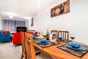 Ein Essbereich mit einem Holztisch, gedeckt für vier Personen, umgeben von Stühlen. Im Hintergrund ist ein Fernseher an der Wand befestigt.