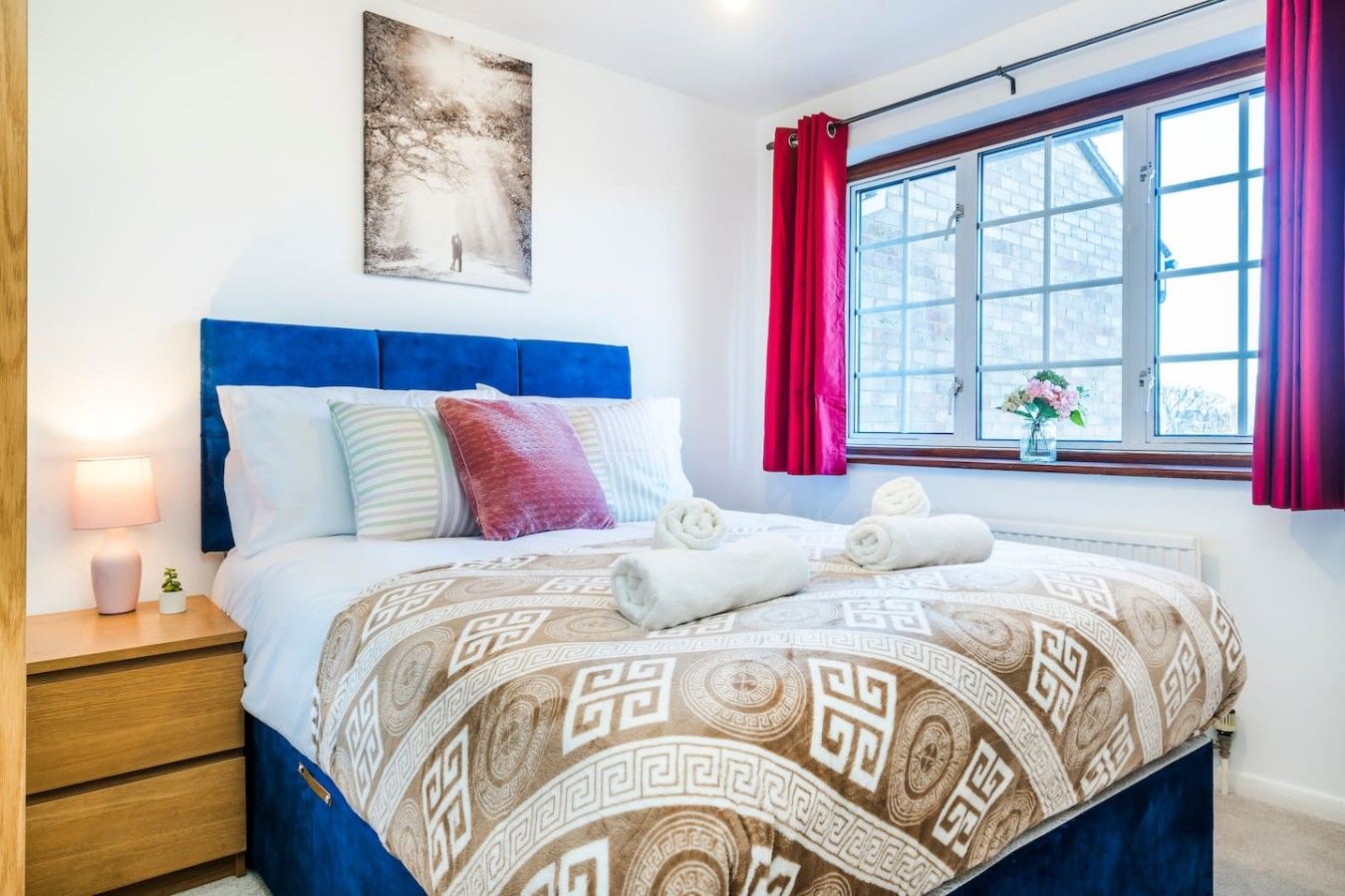 Una cama bien hecha con una colcha estampada, una lámpara de noche y una ventana con cortinas.