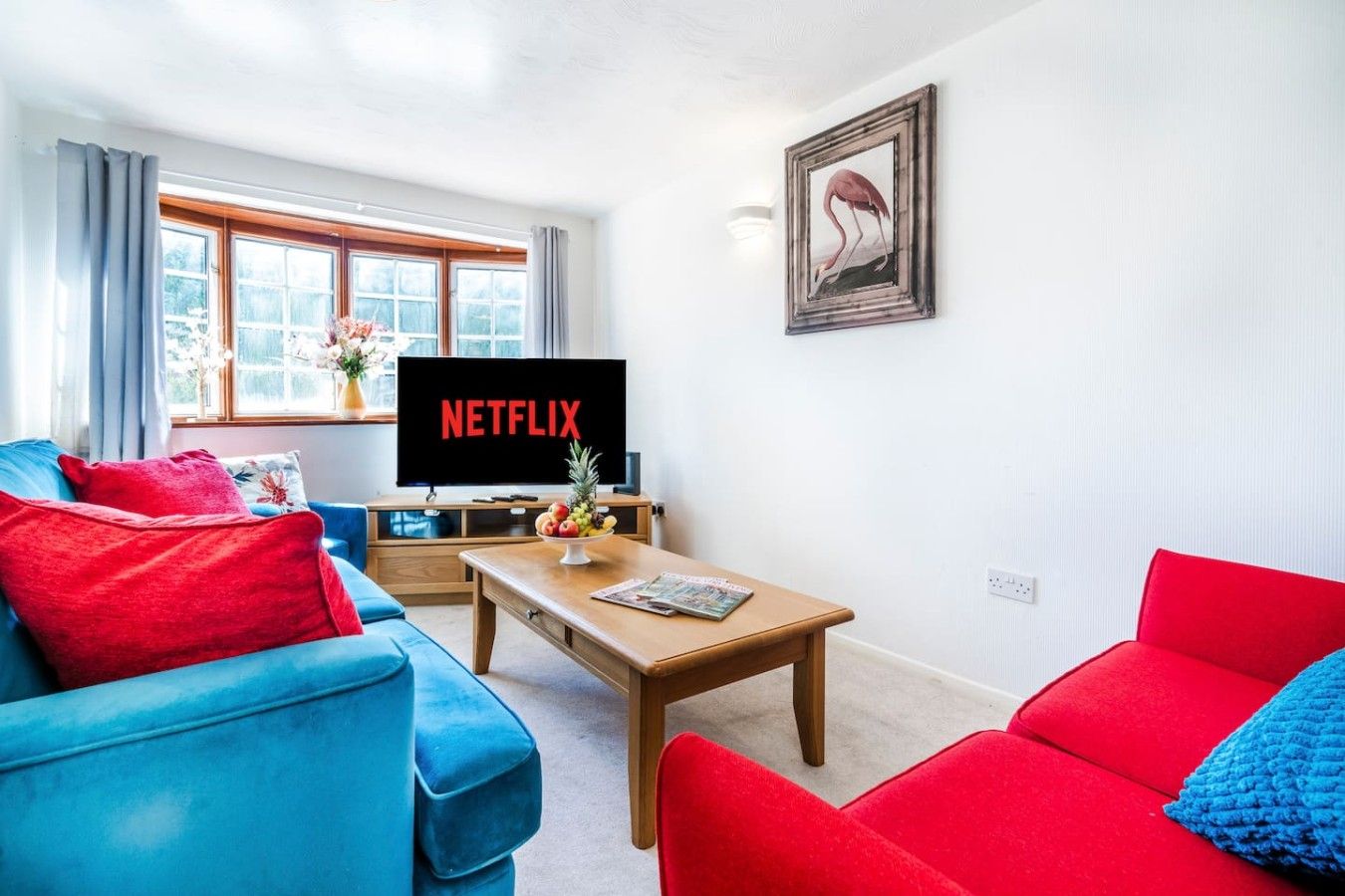 A living room with a television displaying 'NETFLIX,' a coffee table with a fruit bowl, and two sofas in red and blue.