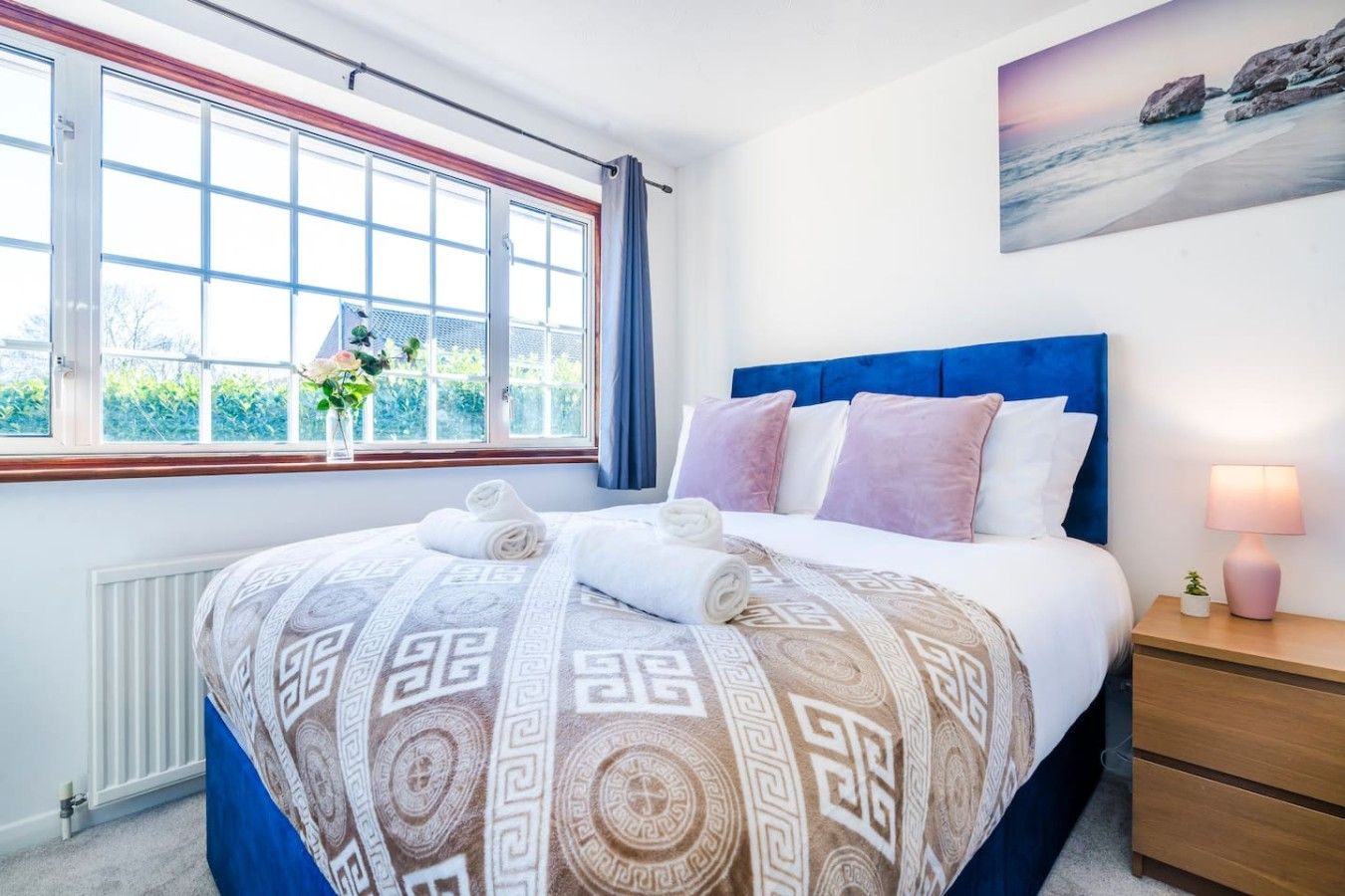 A hotel room with a large bed, a window with curtains, a bedside table with a lamp, and a painting on the wall.