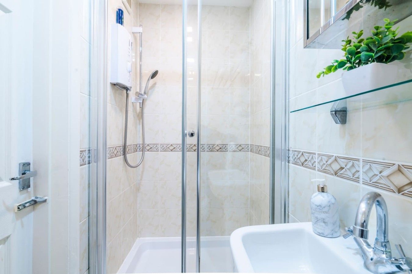 A modern bathroom with a glass-enclosed shower, a white sink with a marble soap dispenser, and a glass shelf with a plant.