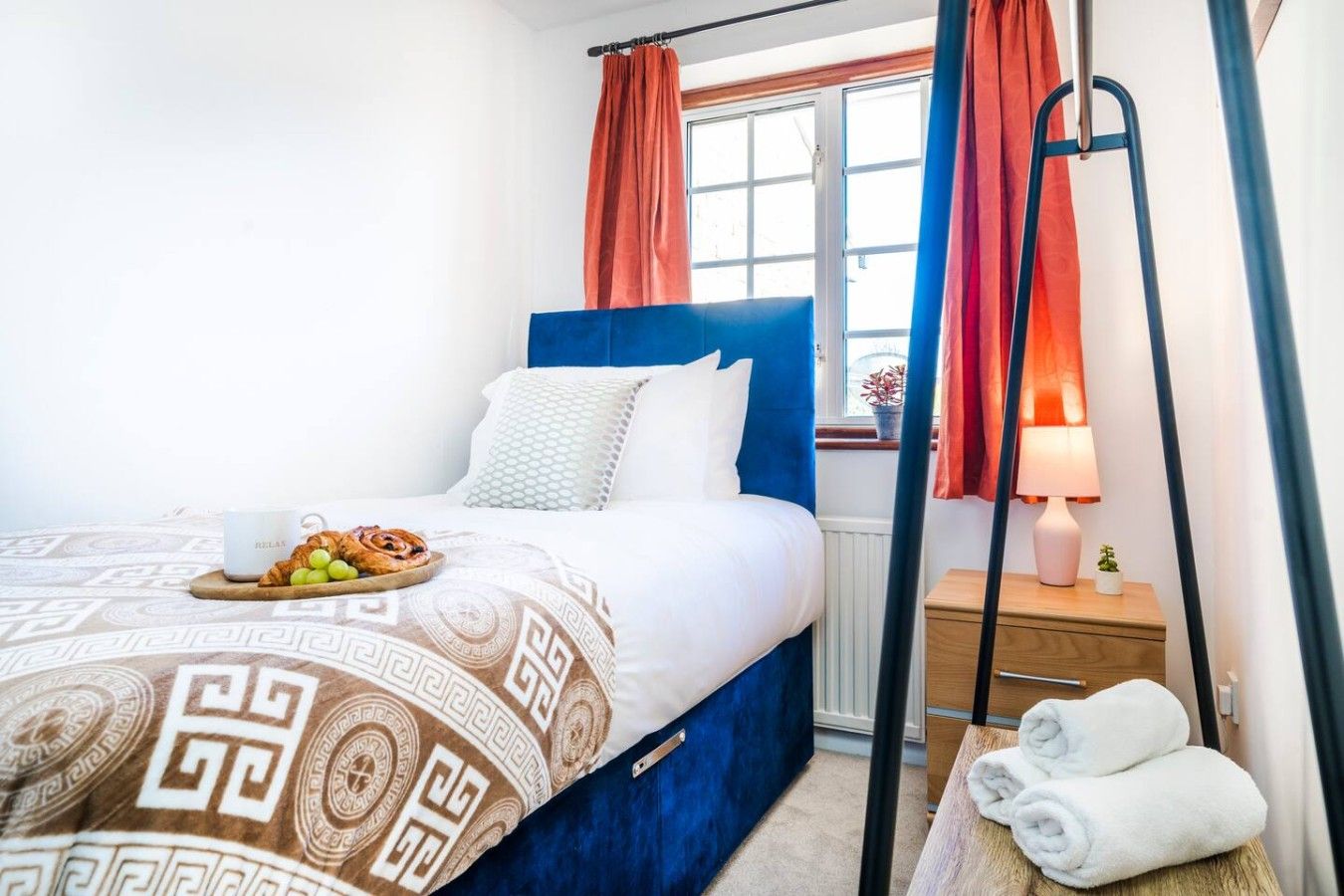 A hotel room with a bed, a bedside table with a lamp, a window with curtains, and a tray with food on the bed.