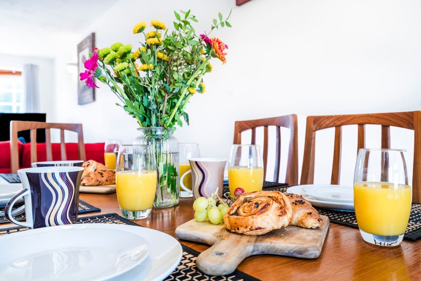 A breakfast setting with a table, plates, glasses of orange juice, a vase with flowers, and a pastry on a cutting board.