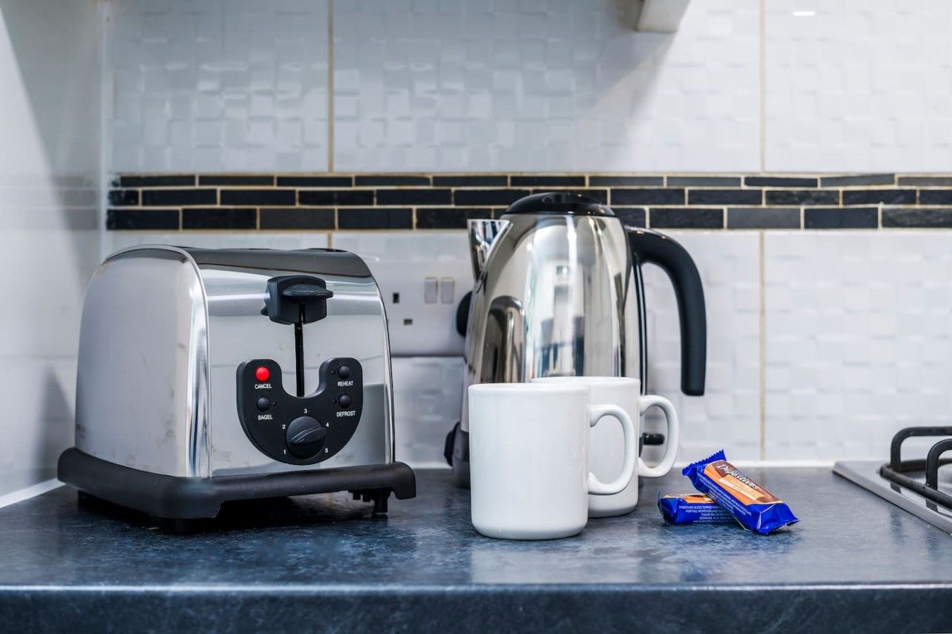 Un plan de travail de cuisine avec un grille-pain, une machine à café, deux tasses blanches et un paquet de sucre.