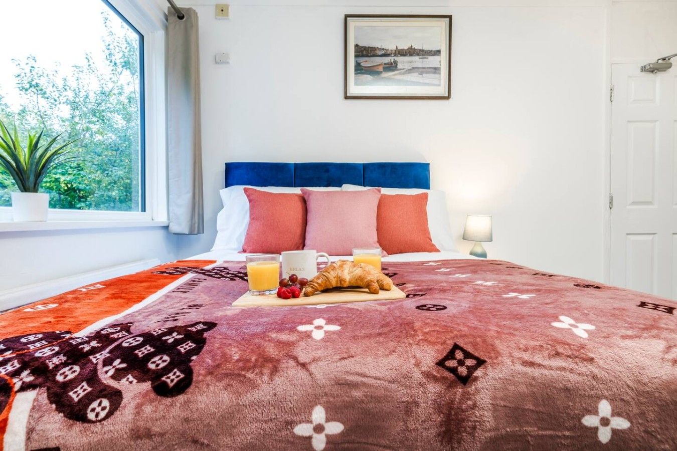 A hotel room with a bed, a window with a view of greenery, a framed picture on the wall, and a bedside table with a lamp.