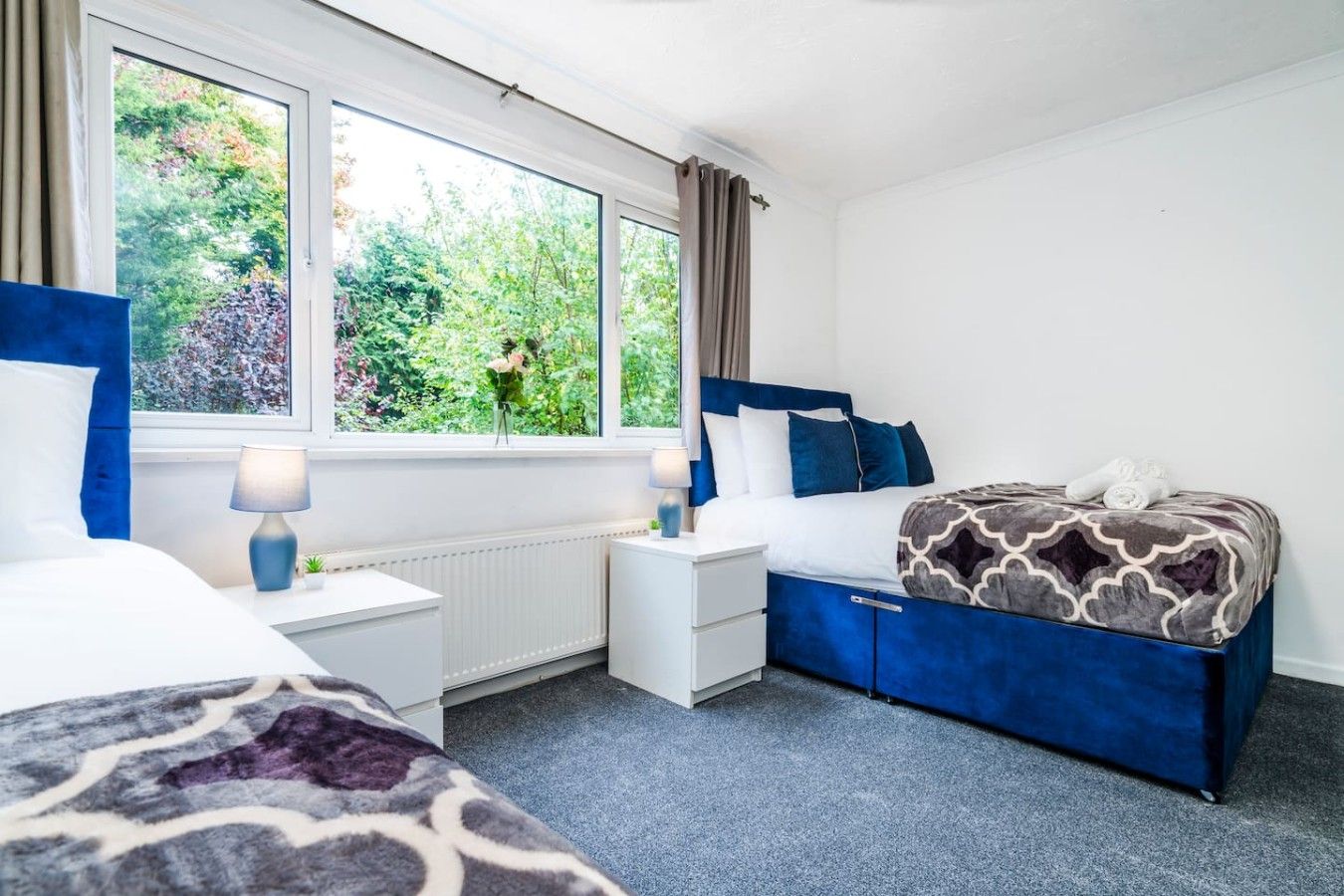 Un dormitorio con cama, mesitas de noche, una ventana con cortinas y vistas a zonas verdes.