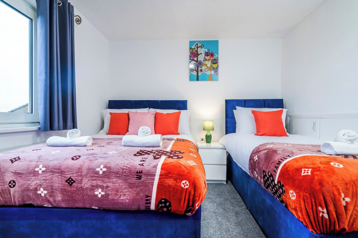 A hotel room with two beds, each with a blue headboard, white bedding, and orange accents. There is a painting on the wall above the beds and a window with curtains on the left.