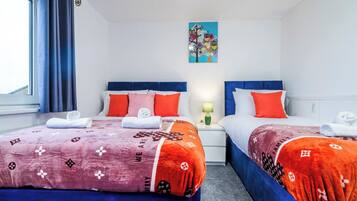 A hotel room with two beds, each with a blue headboard, white bedding, and orange accents. There is a painting on the wall above the beds and a window with curtains on the left.