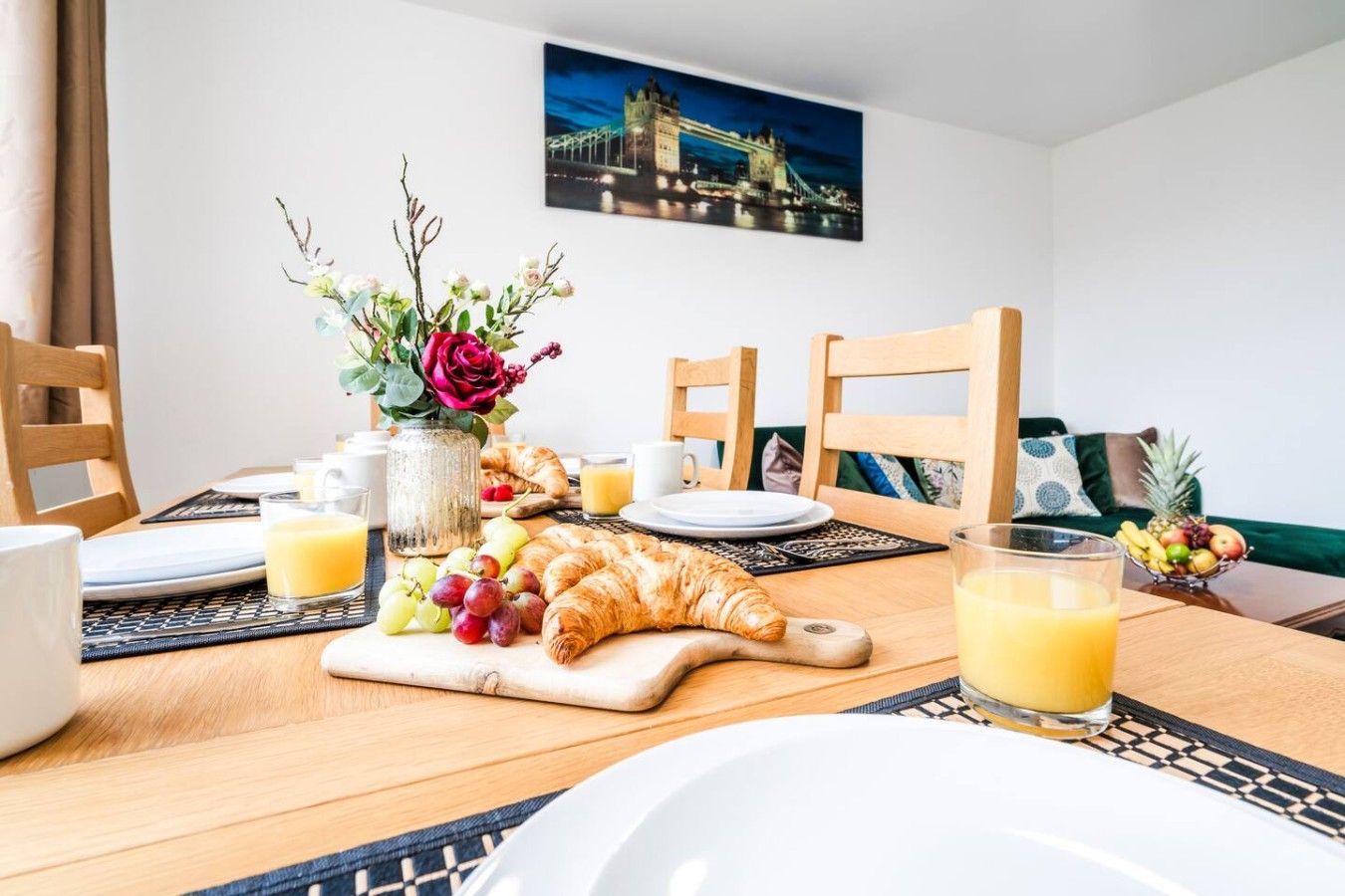 A breakfast setting with croissants, grapes, and juice on a wooden table, with chairs and a vase of flowers.