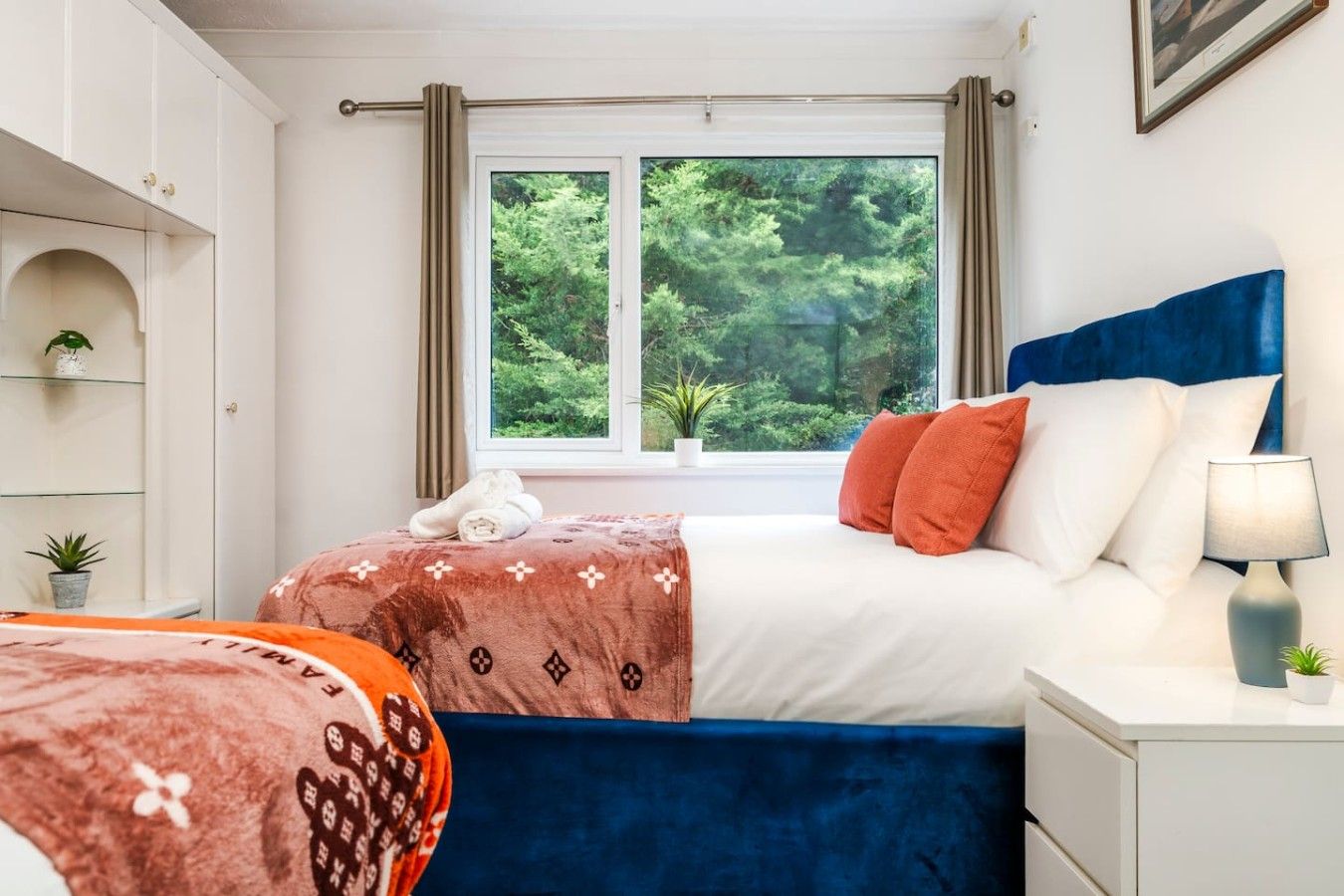 Un dormitorio con cama, mesita de noche, ventana con cortinas, una planta y una estantería con una planta.