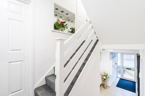Una escalera interior blanca con barandilla de cristal, que lleva a un rellano con una puerta y una ventana.