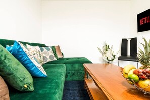 Ein grünes Sofa mit gemusterten Kissen, ein Holztisch mit Obst und ein an der Wand befestigtes Netflix-Schild.