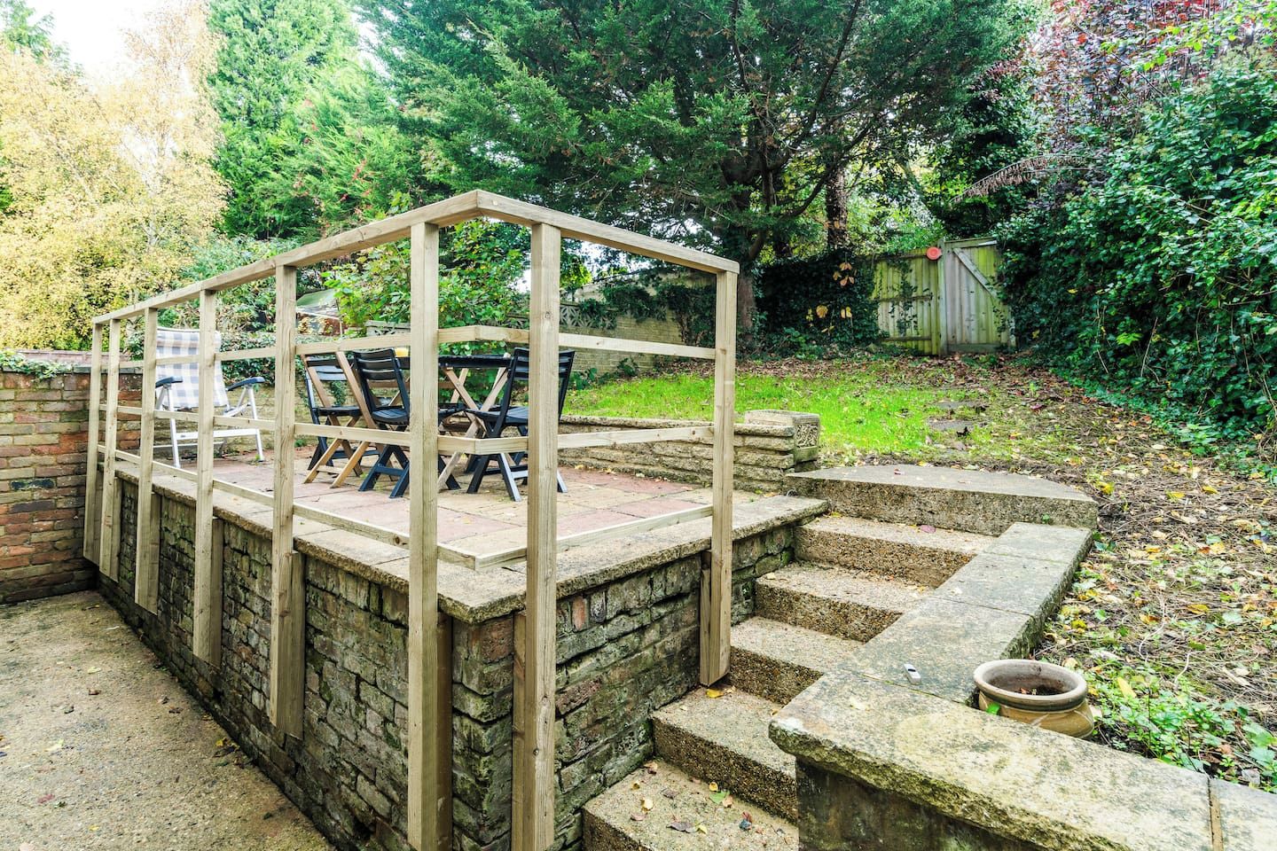 A wooden pergola structure with a table and chairs underneath, surrounded by a stone wall and steps leading to a garden area.