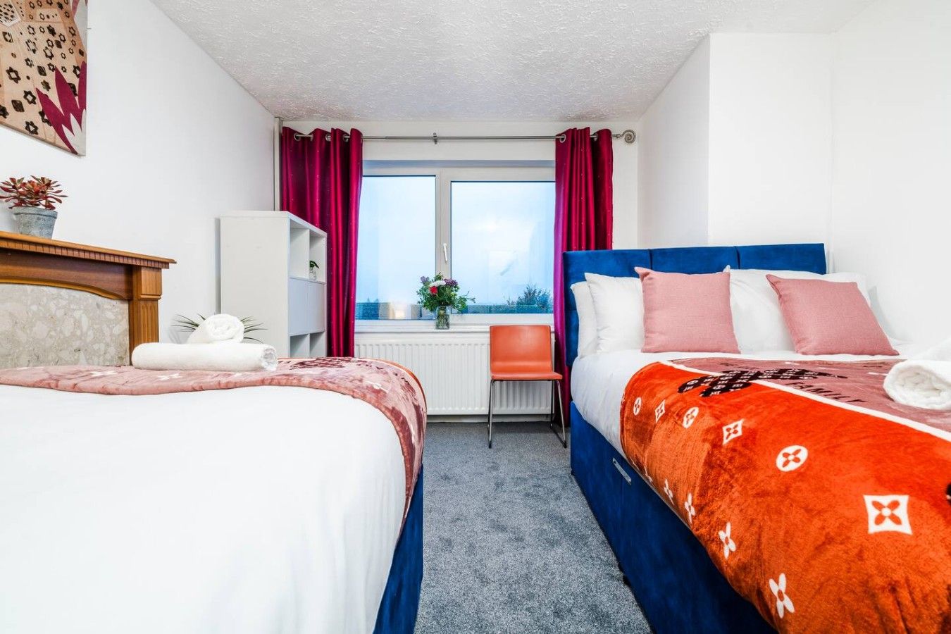Une chambre d’hôtel avec deux lits, une fenêtre avec des rideaux, un radiateur, une chaise et une petite table sur laquelle est posée une plante.