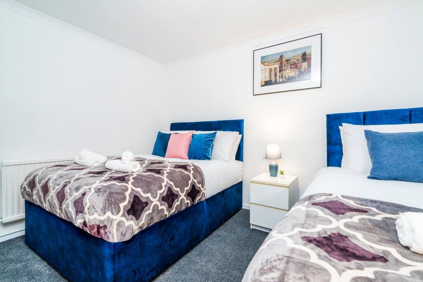 A hotel room with two beds, a nightstand with a lamp, and a framed picture on the wall.
