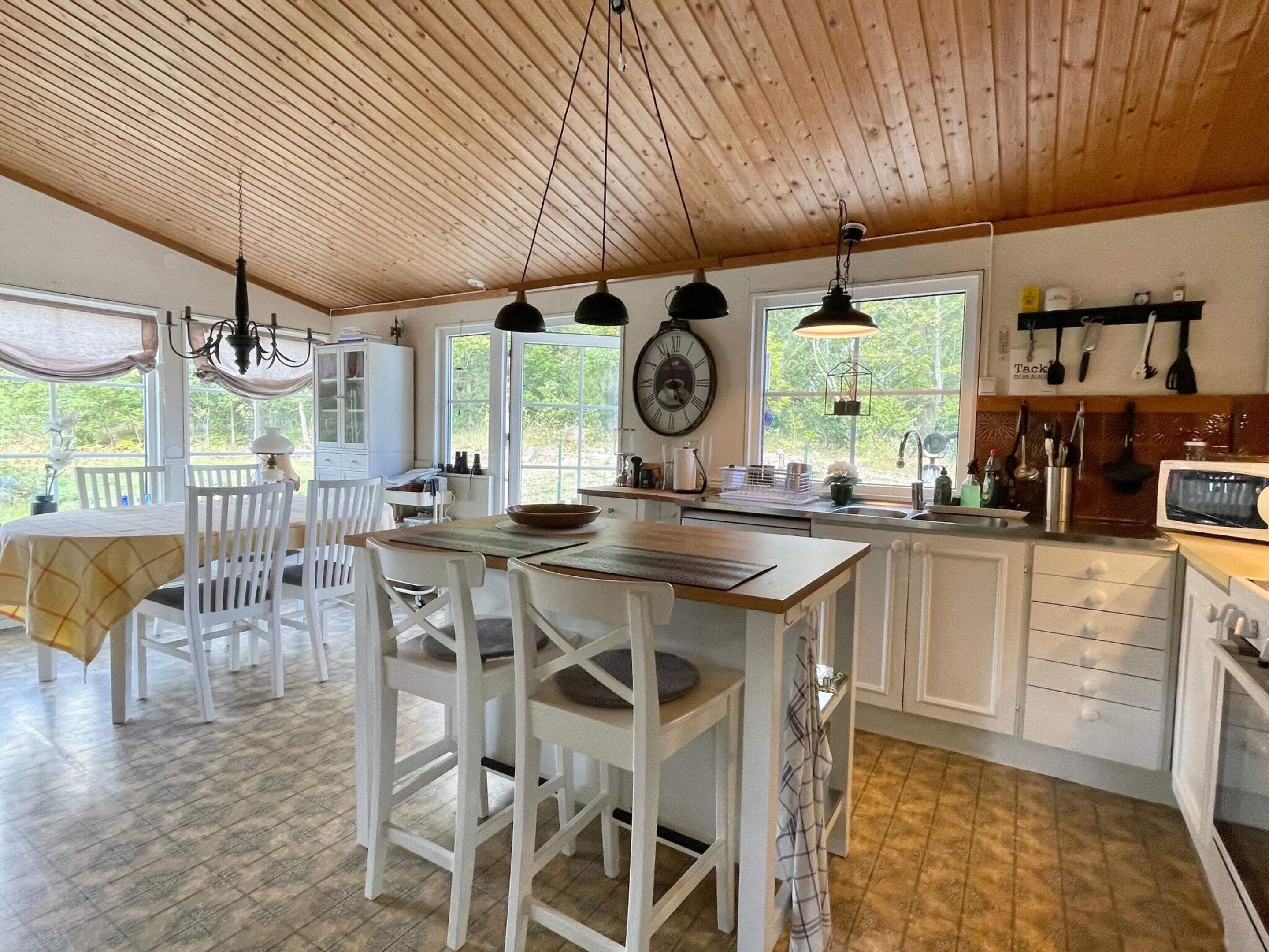 A kitchen with wooden ceiling, white cabinets, a dining area with white chairs, and a view of greenery outside.