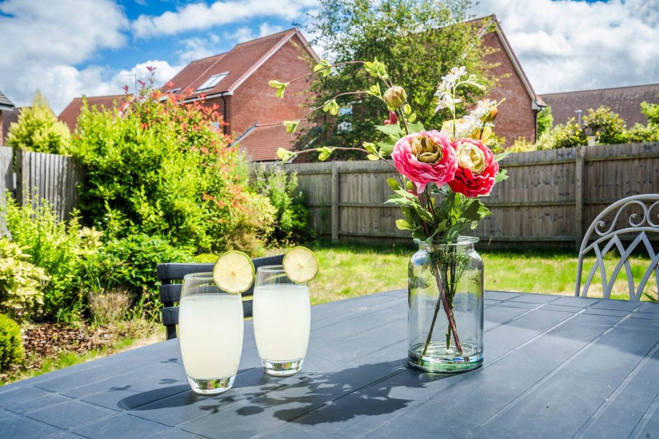 A table with two glasses of a clear beverage, a vase with pink flowers, and a wooden fence in the background.
