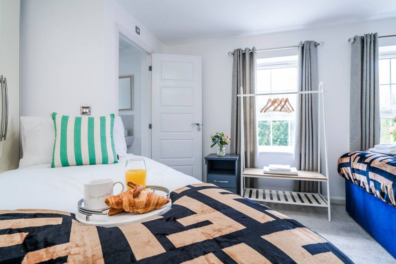 Ett hotellrum med en säng, ett nattduksbord med ett glas apelsinjuice och en croissant, ett fönster med gardiner och utsikt över grönområden utanför.