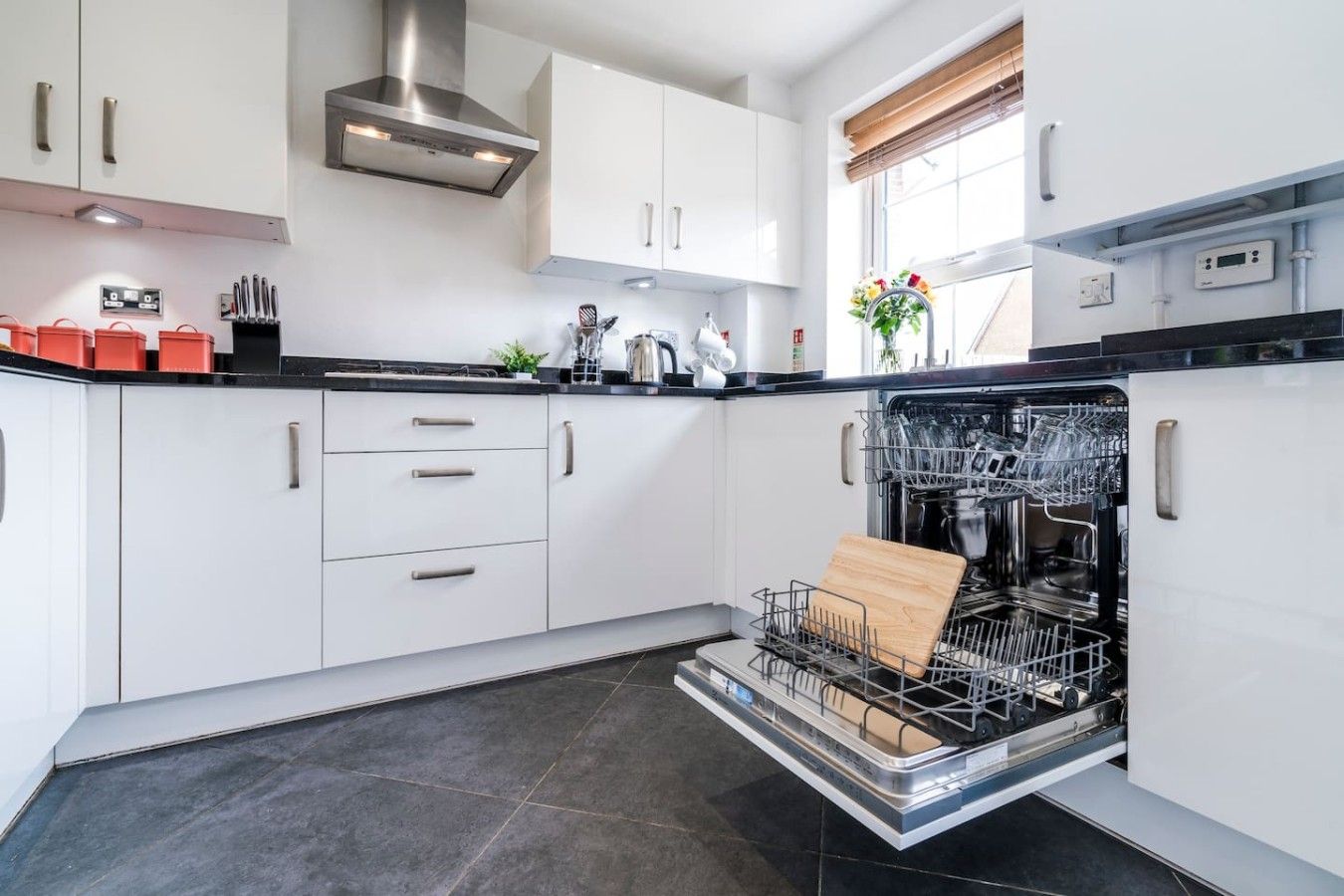 Una cocina moderna con armarios blancos, extractor de acero inoxidable y lavavajillas con tabla de cortar en su interior.